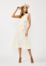 [Color: Natural] A front facing image of a blonde model wearing an off white sleeveless shirt dress with a collared neckline, button front, and a self tie waist belt. 