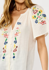 [Color: Cream] A detail image of a blonde model wearing a light sheer cotton top with floral embroidery. Featuring a short dolman sleeves, a split v-neckline, and button front. 