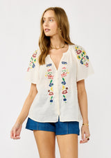 [Color: Cream] A front facing image of a blonde model wearing a light sheer cotton top with floral embroidery. Featuring a short dolman sleeves, a split v-neckline, and button front. 