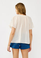 [Color: Cream] A back facing image of a blonde model wearing a light sheer cotton top with floral embroidery. Featuring a short dolman sleeves, a split v-neckline, and button front. 