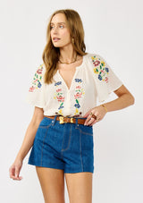 [Color: Cream] A front facing image of a blonde model wearing a light sheer cotton top with floral embroidery. Featuring a short dolman sleeves, a split v-neckline, and button front. 