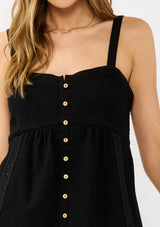 [Color: Black] A detail image of a blonde model wearing a black boho tank top with a smocked back, button front, an eyelet trim, and pintuck details. Perfect for everyday warm weather styling. 