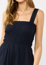 [Color: Navy] A detail image of a blonde model wearing a navy wide leg jumpsuit with a smocked top, wide fixed straps, and side pockets. 