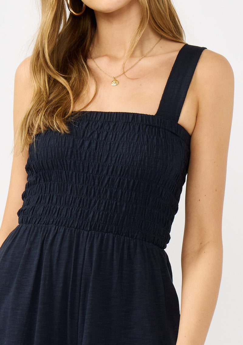[Color: Navy] A detail image of a blonde model wearing a navy wide leg jumpsuit with a smocked top, wide fixed straps, and side pockets. 