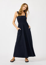 [Color: Navy] A front facing image of a blonde model wearing a navy wide leg jumpsuit with a smocked top, wide fixed straps, and side pockets. 