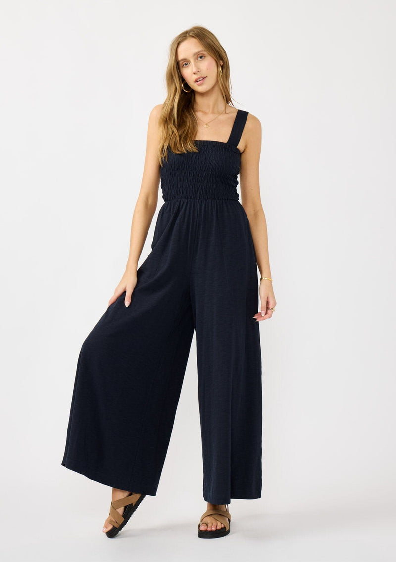 [Color: Navy] A front facing image of a blonde model wearing a navy wide leg jumpsuit with a smocked top, wide fixed straps, and side pockets. 