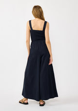[Color: Navy] A back facing image of a blonde model wearing a navy wide leg jumpsuit with a smocked top, wide fixed straps, and side pockets. 
