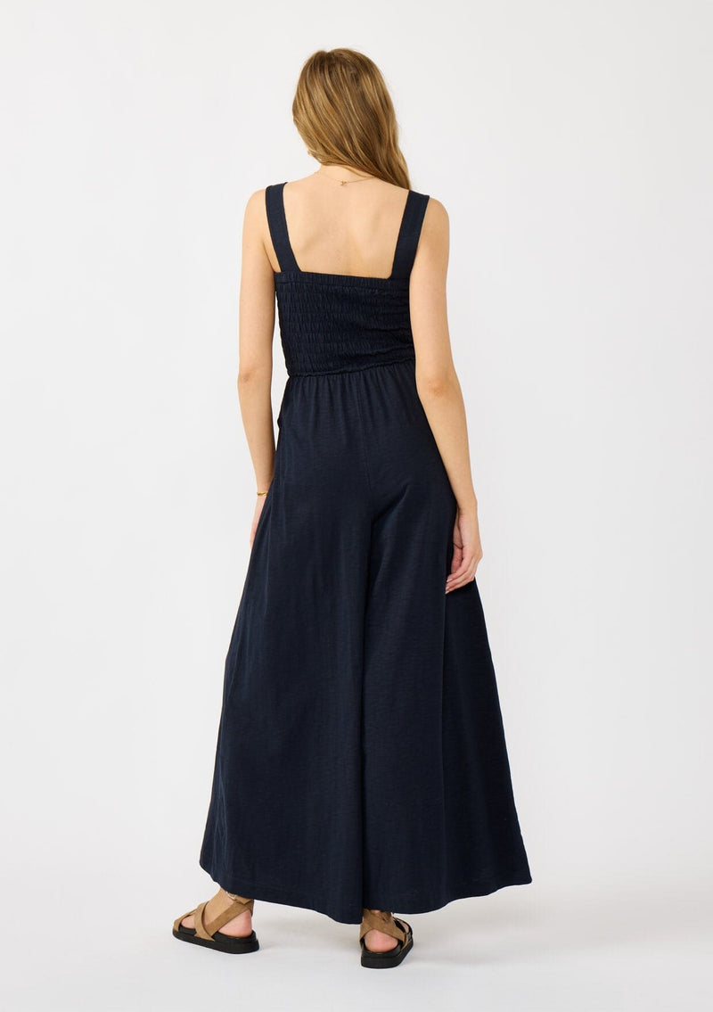 [Color: Navy] A back facing image of a blonde model wearing a navy wide leg jumpsuit with a smocked top, wide fixed straps, and side pockets. 