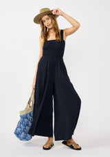 [Color: Navy] A front facing image of a blonde model wearing a navy wide leg boho jumpsuit with a smocked top, wide fixed straps, and side pockets. Styled with a boho bag and hat. 