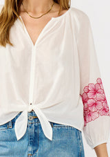 [Color: Off White/Fuchsia] A detail image of a blonde model wearing a white sheer tie-front top, featuring delicate pink floral embroidered cutouts along the sleeves.