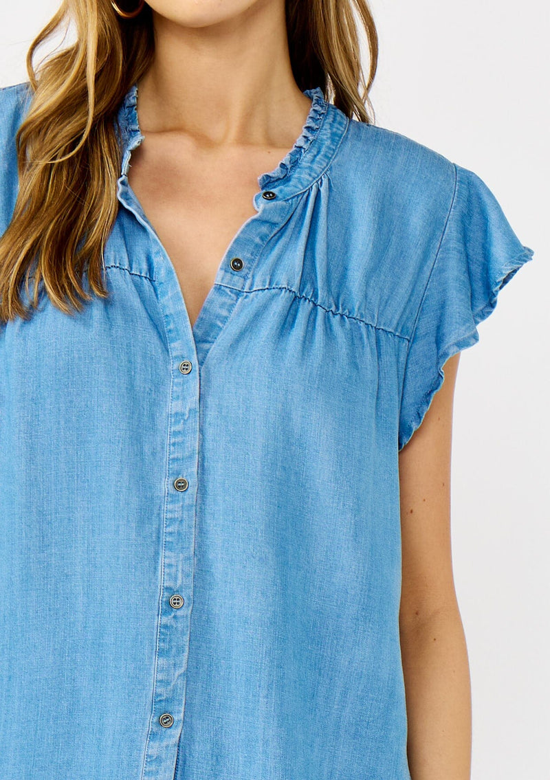 [Color: Light Denim Blue] A detail image of a blonde model wearing a soft blue denim Tencel top featuring a button front, flutter sleeves, and ruffle trim accents. A lightweight and flowy summer top.