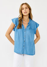 [Color: Light Denim Blue] A front facing image of a blonde model wearing a soft blue denim Tencel top featuring a button front, flutter sleeves, and ruffle trim accents. A lightweight and flowy summer top.