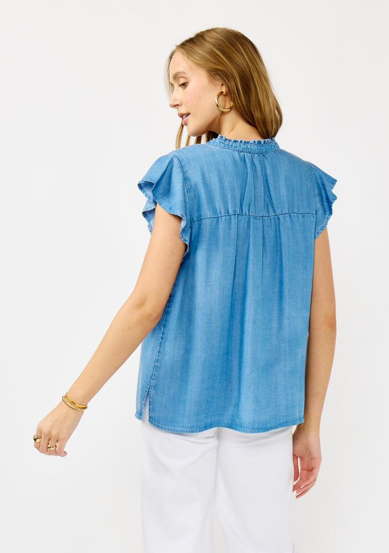 [Color: Light Denim Blue] A back facing image of a blonde model wearing a soft blue denim Tencel top featuring a button front, flutter sleeves, and ruffle trim accents. A lightweight and flowy summer top.