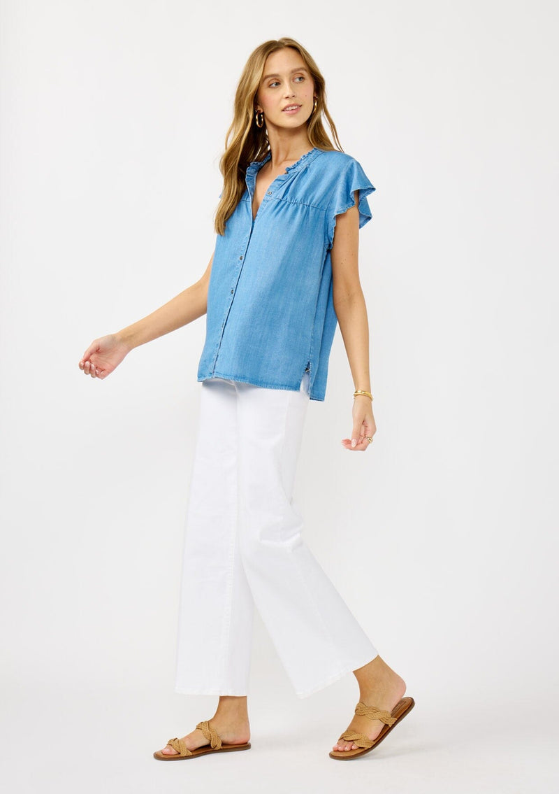 [Color: Light Denim Blue] A side facing image of a blonde model wearing a soft blue denim Tencel top featuring a button front, flutter sleeves, and ruffle trim accents. A lightweight and flowy summer top.