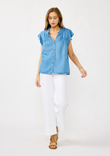 [Color: Light Denim Blue] A front facing image of a blonde model wearing a soft blue denim Tencel top featuring a button front, flutter sleeves, and ruffle trim accents. A lightweight and flowy summer top.