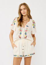 [Color: Cream] A front facing image of a blonde model wearing cream floral embroidered short crafted in soft cotton with  matching top. Featuring a smocked waist, a drawstring, and side pockets. 