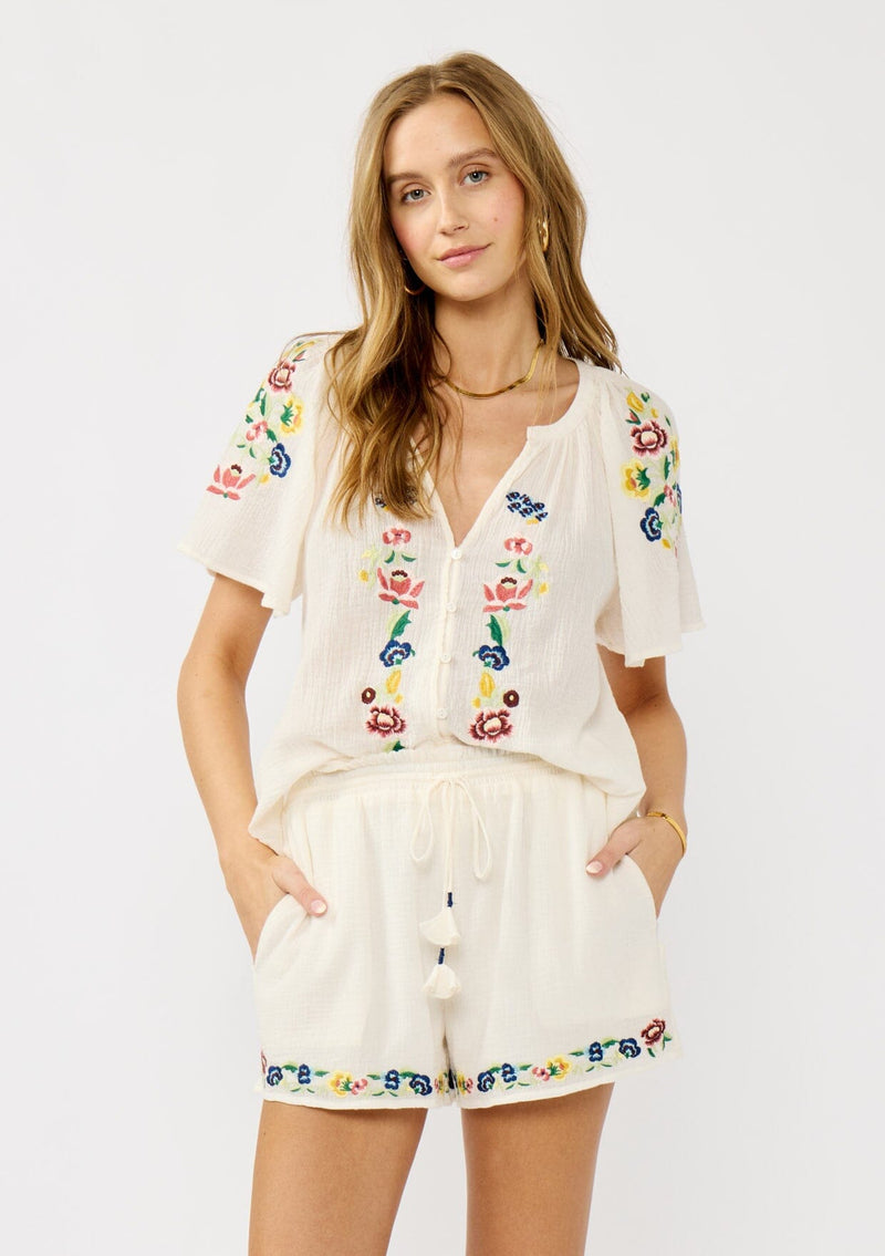[Color: Cream] A front facing image of a blonde model wearing cream floral embroidered short crafted in soft cotton with  matching top. Featuring a smocked waist, a drawstring, and side pockets. 