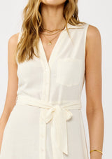 [Color: Natural] A detail image of a blonde model wearing an off white sleeveless shirt dress with a collared neckline, button front, and a self tie waist belt. 