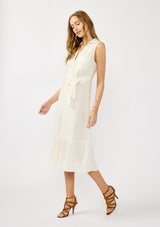 [Color: Natural] A side facing image of a blonde model wearing an off white sleeveless shirt dress with a collared neckline, button front, and a self tie waist belt. 