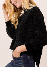 [Color: Black] A blonde model wearing a cozy cable knit sweater made with super soft chenille. Featuring a flattering ribbed funnel neck and a chic high low hemline with side vents. 