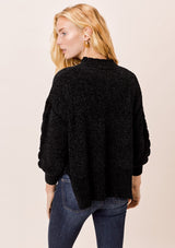 [Color: Black] A blonde model wearing a cozy cable knit sweater made with super soft chenille. Featuring a flattering ribbed funnel neck and a chic high low hemline with side vents. 