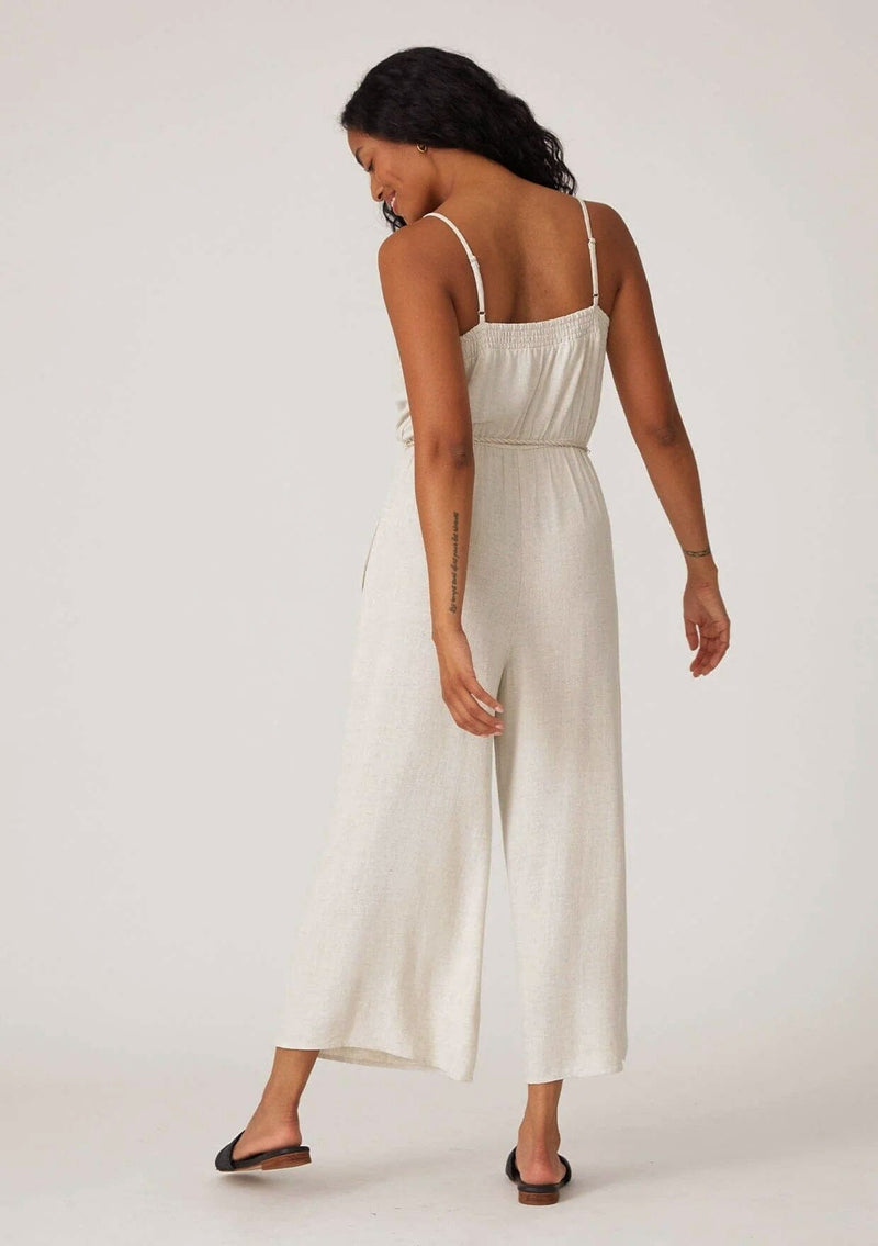 [Color: Natural] A back facing image of a brunette model wearing an off white sleeveless jumpsuit. With adjustable spaghetti straps, a scooped neckline, a long wide leg, side pockets, an elastic waist, and a braded tassel belt.