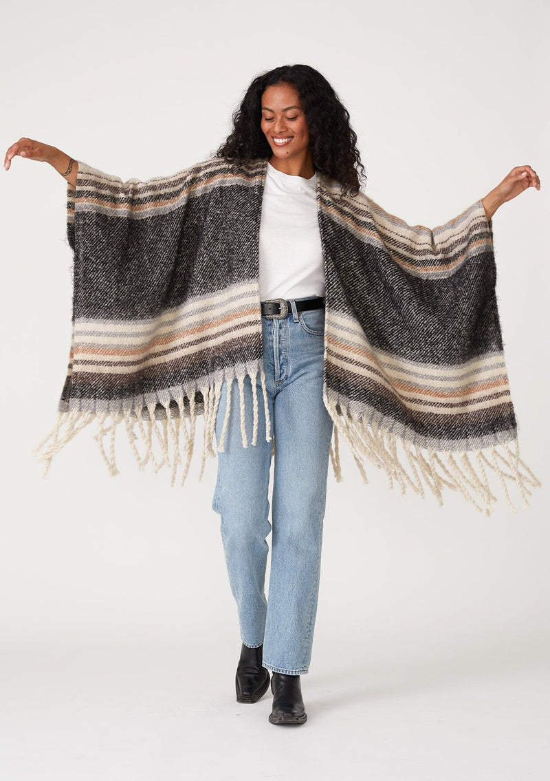 [Color: Navy/Taupe] A full body front facing image of a brunette model wearing a soft sweater shawl in a navy blue and taupe stripe. With a fringed hemline, a mid length silhouette, an open front, and long sleeves. 
