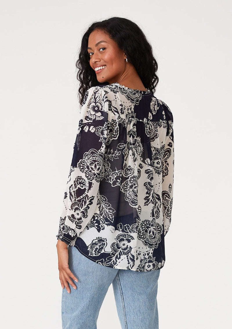 [Color: Natural/Navy] A back facing image of a brunette model wearing a flowy chiffon bohemian blouse in a blue and white floral print. With voluminous long sleeves, ruffle details, and a v neckline with doubles ties. 