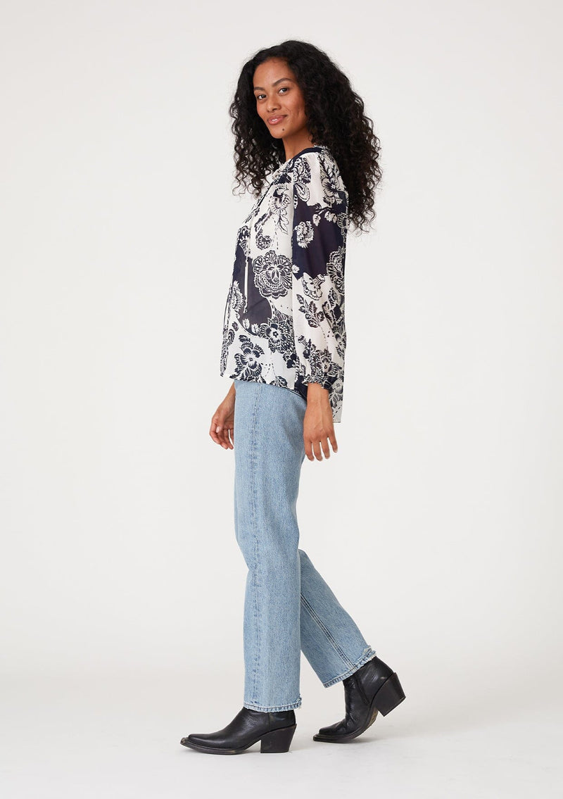 [Color: Natural/Navy] A side facing image of a brunette model wearing a flowy chiffon bohemian blouse in a blue and white floral print. With voluminous long sleeves, ruffle details, and a v neckline with doubles ties. 