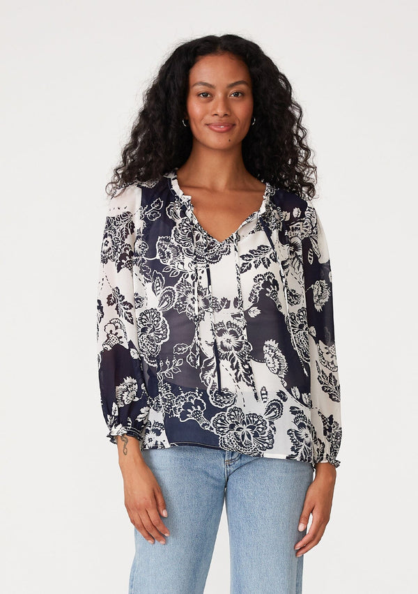 [Color: Natural/Navy] A front facing image of a brunette model wearing a flowy chiffon bohemian blouse in a blue and white floral print. With voluminous long sleeves, ruffle details, and a v neckline with doubles ties. 