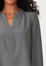 [Color: Moss] A close up front facing image of a brunette model wearing a moss green blouse with a v neckline, long sleeves, a relaxed fit, and a smocked detail at the back.