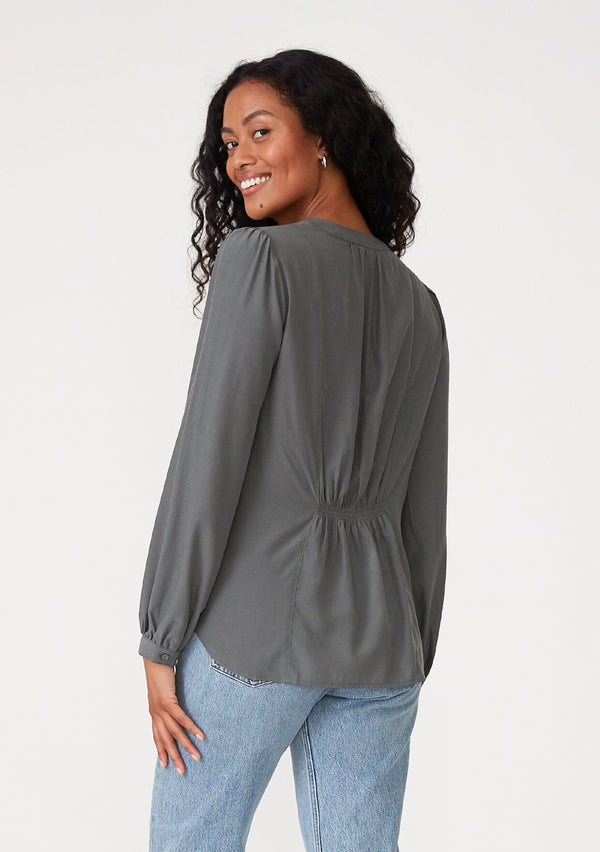 [Color: Moss] A back facing image of a brunette model wearing a moss green blouse with a v neckline, long sleeves, a relaxed fit, and a smocked detail at the back.