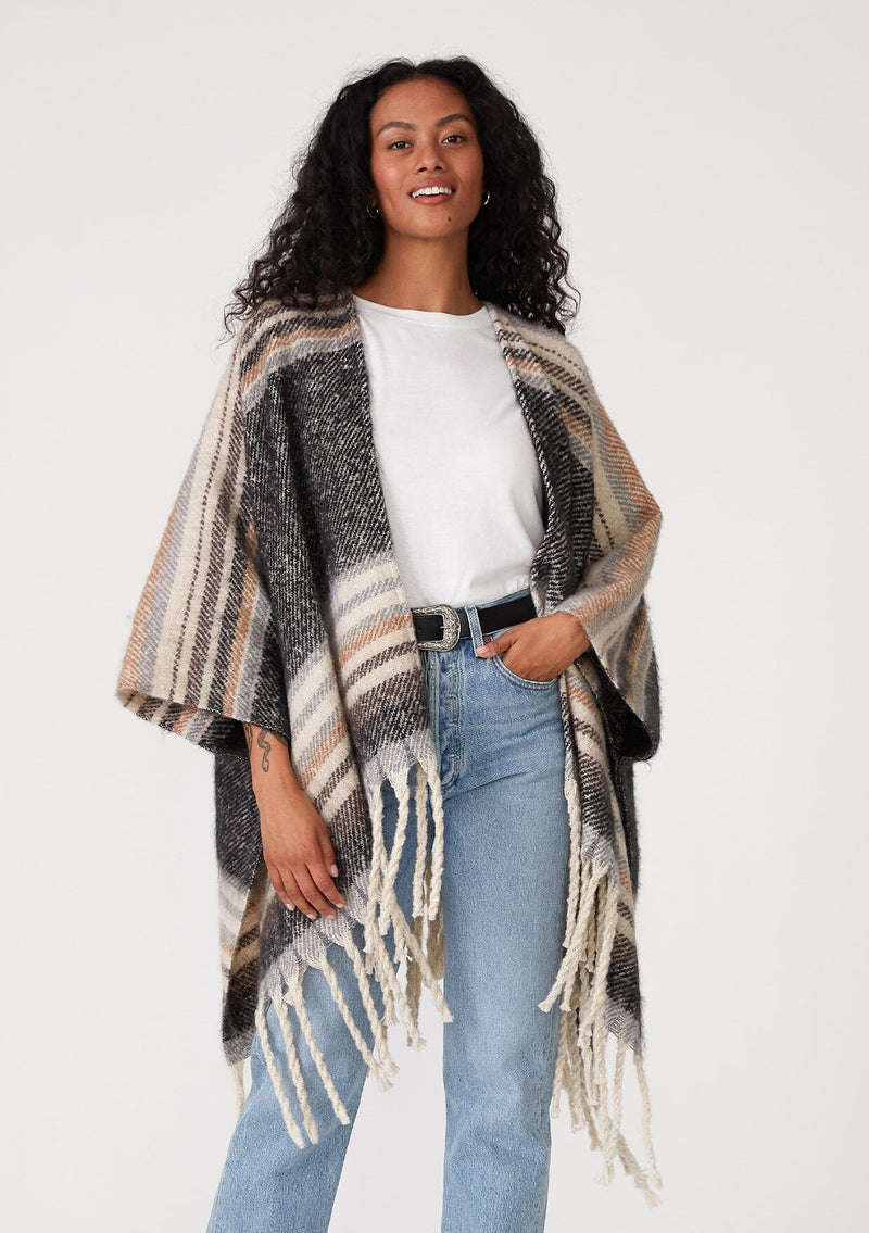 [Color: Navy/Taupe] A front facing image of a brunette model wearing a soft sweater shawl in a navy blue and taupe stripe. With a fringed hemline, a mid length silhouette, an open front, and long sleeves. 