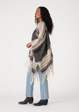 [Color: Navy/Taupe] A side facing image of a brunette model wearing a soft sweater shawl in a navy blue and taupe stripe. With a fringed hemline, a mid length silhouette, an open front, and long sleeves. 