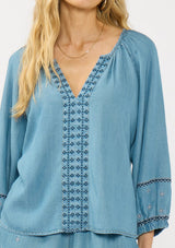[Color: Vintage Blue] A detail image of a blonde model wearing a vintage blue blouse with embroidered details, a split v neckline, and elastic cuffs. Styled with matching boho embroidered shorts. 