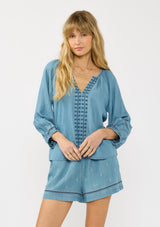 [Color: Vintage Blue] A front facing image of a blonde model wearing a vintage blue blouse with embroidered details, a split v neckline, and elastic cuffs. Styled with matching boho embroidered shorts. 