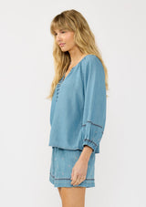 [Color: Vintage Blue] A side facing image of a blonde model wearing a vintage blue blouse with embroidered details, a split v neckline, and elastic cuffs. Styled with matching boho embroidered shorts. 