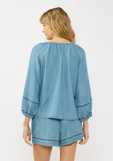 [Color: Vintage Blue] A back facing image of a blonde model wearing a vintage blue blouse with embroidered details, a split v neckline, and elastic cuffs. Styled with matching boho embroidered shorts. 