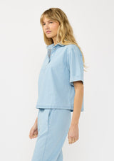 [Color: Light Denim Blue] A side facing image of  blonde model wearing a cotton short sleeve shirt with a half button front and a collared neck. Styled with matching bottoms. 