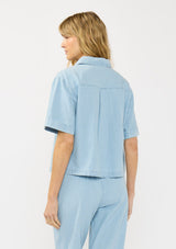 [Color: Light Denim Blue] A back facing image of  blonde model wearing a cotton short sleeve shirt with a half button front and a collared neck. Styled with matching bottoms. 