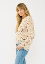 [Color: Natural] A side facing image of a blonde model wearing a circular crochet knit top with a round neckline and long sleeves. A spring boho top for casual and beach styling. 