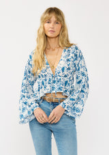 [Color: Ivory/Navy] A front facing image of a blonde model wearing a blue and white floral top with a tie front, deep v neckline, ruffle trimmed hem, and long flowy sleeves.