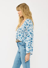 [Color: Ivory/Navy] A side facing image of a blonde model wearing a blue and white floral top with a tie front, deep v neckline, ruffle trimmed hem, and long flowy sleeves.