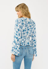 [Color: Ivory/Navy] A back facing image of a blonde model wearing a blue and white floral top with a tie front, deep v neckline, ruffle trimmed hem, and long flowy sleeves.