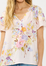 [Color: Blush/Dusty Blue] A detail image of a blonde model wearing a pink floral top with layered short sleeves and a split v-neckline. A spring feminine top for casual outings. 