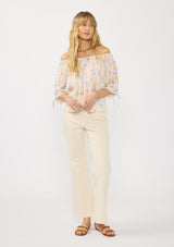 [Color: Almond/Rose] A front facing image of a blonde model wearing a colorful bohemian off shoulder top with an ikat print, a smocked elastic neckline and long sleeves with a ruched detail. 