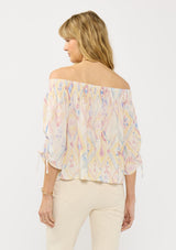 [Color: Almond/Rose] A back facing image of a blonde model wearing a colorful bohemian off shoulder top with an ikat print, a smocked elastic neckline and long sleeves with a ruched detail. 