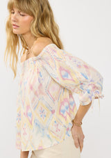 [Color: Almond/Rose] A side facing image of a blonde model wearing a colorful bohemian off shoulder top with an ikat print, a smocked elastic neckline and long sleeves with a ruched detail. 