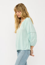 [Color: Dusty Mint] A side facing image of a blonde model wearing a mint green boho blouse with embroidered and lace details. 