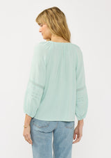 [Color: Dusty Mint] A back facing image of a blonde model wearing a mint green boho blouse with embroidered and lace details. 